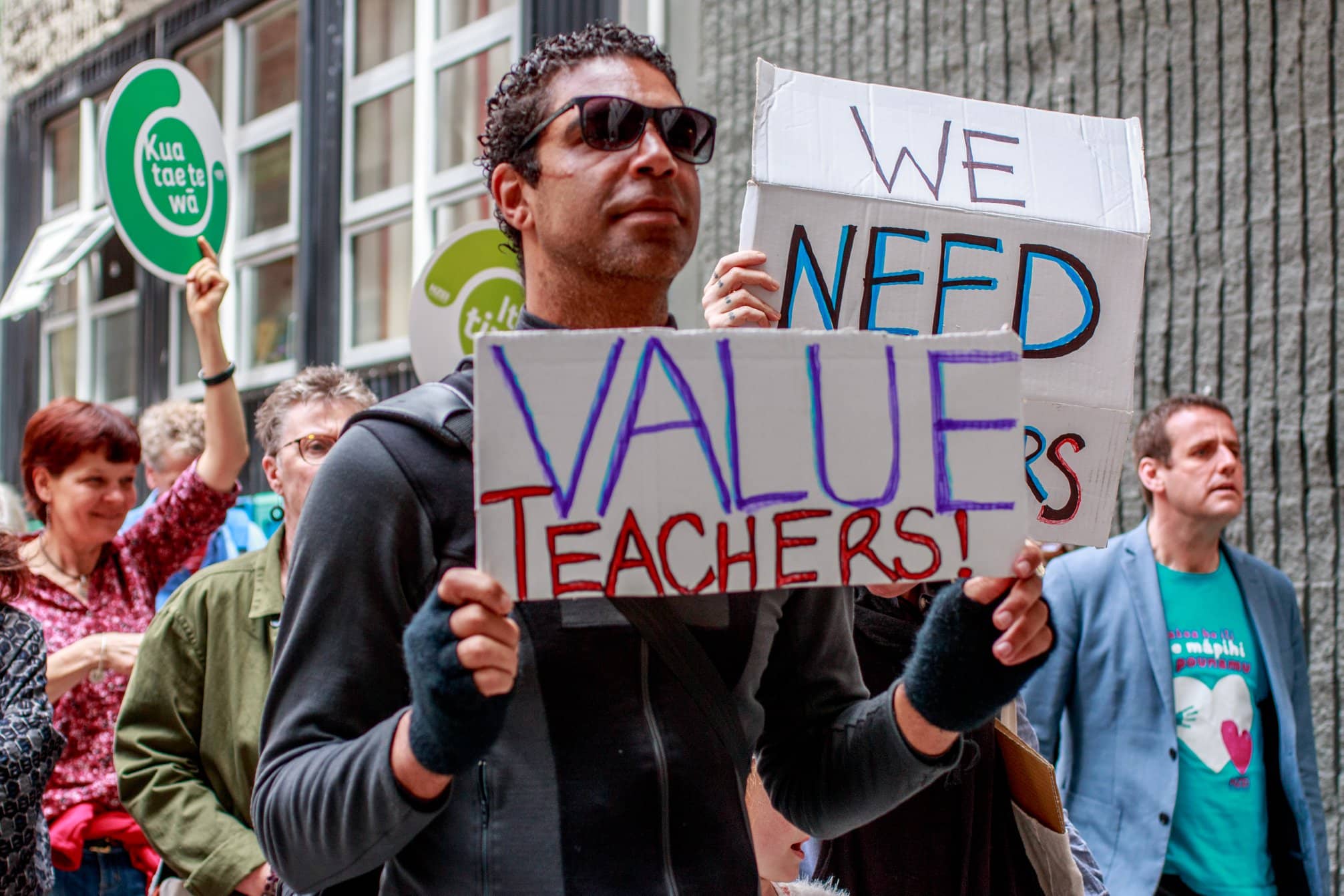People march down the streets of Wellington, and we see the close up of several dedicated volunteers or teachers raising signs, “Value teachers”, and “We need teachers”